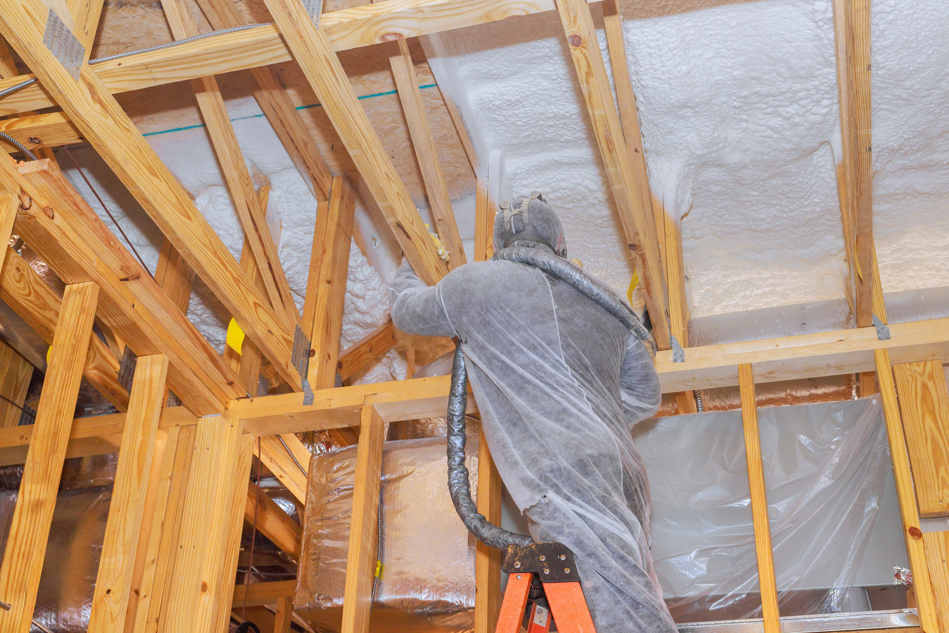 Construction worker in protective suit on a ladder amid exposed wooden framing and white spray foam insulation in a construction site gemeral area.